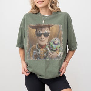 May include: An olive green short-sleeve t-shirt featuring a graphic print of the characters Woody and Buzz Lightyear. Woody is wearing dark sunglasses. The graphic has a distressed, vintage-style square border on the front of the shirt.