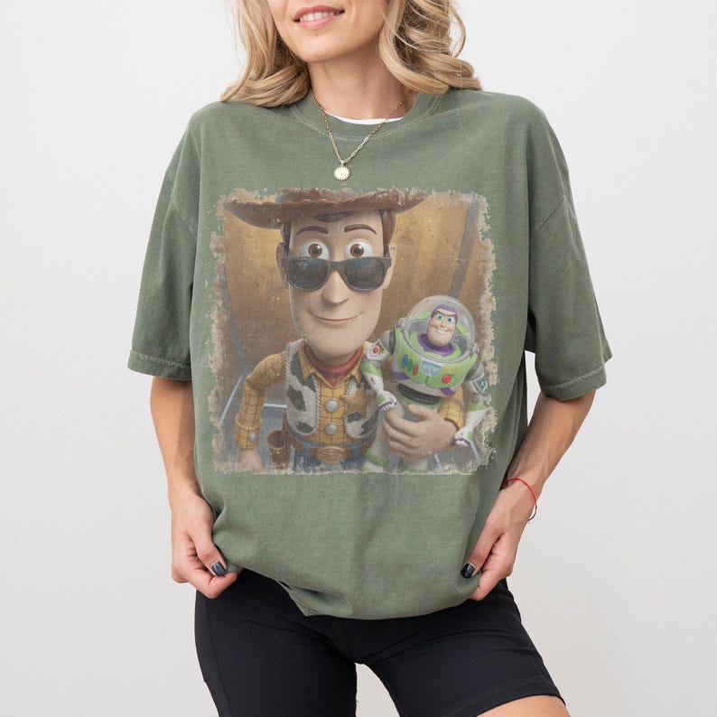 May include: An olive green short-sleeve t-shirt featuring a graphic print of the characters Woody and Buzz Lightyear. Woody is wearing dark sunglasses. The graphic has a distressed, vintage-style square border on the front of the shirt.