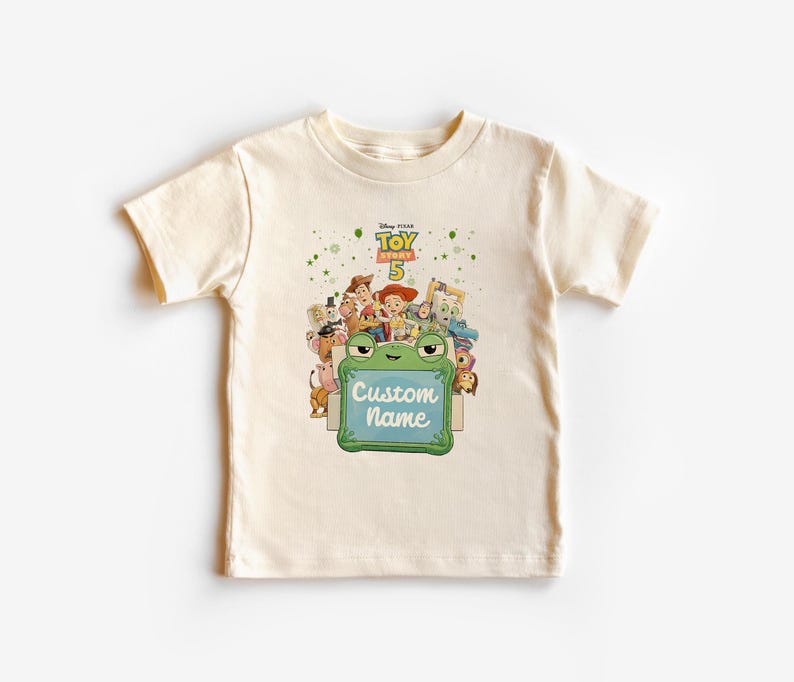 Puede incluir: Una camiseta color crema con un gr&aacute;fico colorido de personajes de la pel&iacute;cula Disney Pixar Toy Story 5. El gr&aacute;fico incluye el texto "Custom Name" en un letrero verde con forma de rana.