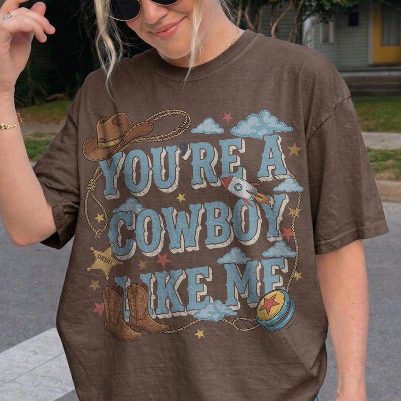 Puede incluir: Una camiseta marr&oacute;n de manga corta con el texto "YOU'RE A COWBOY LIKE ME" en azul claro. El gr&aacute;fico incluye un sombrero de vaquero marr&oacute;n, un lazo, una estrella de sheriff, un cohete, botas de vaquero marrones, un yo-yo azul y estrellas.