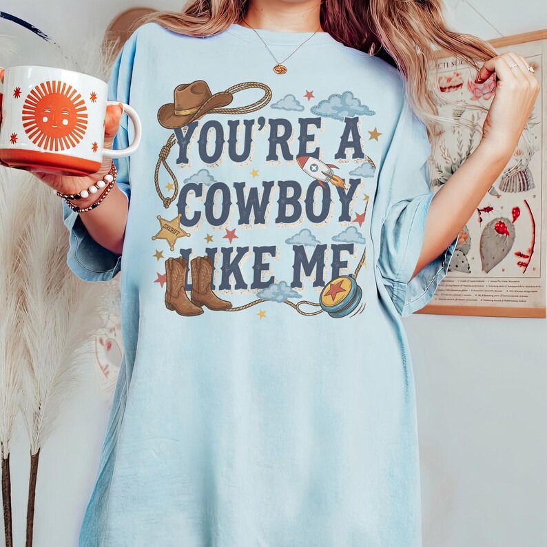 Puede incluir: Una camiseta azul claro con un estampado gr&aacute;fico que incluye el texto "YOU'RE A COWBOY LIKE ME" en letras azul oscuro. El dise&ntilde;o presenta un sombrero de vaquero marr&oacute;n, un lazo, una estrella de sheriff, botas marrones, un cohete y un yo-yo.
