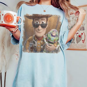 May include: A light blue t-shirt featuring a graphic print of Woody wearing sunglasses and holding Buzz Lightyear. The shirt is paired with a white mug decorated with a large orange sun design and a beaded bracelet on the wrist.