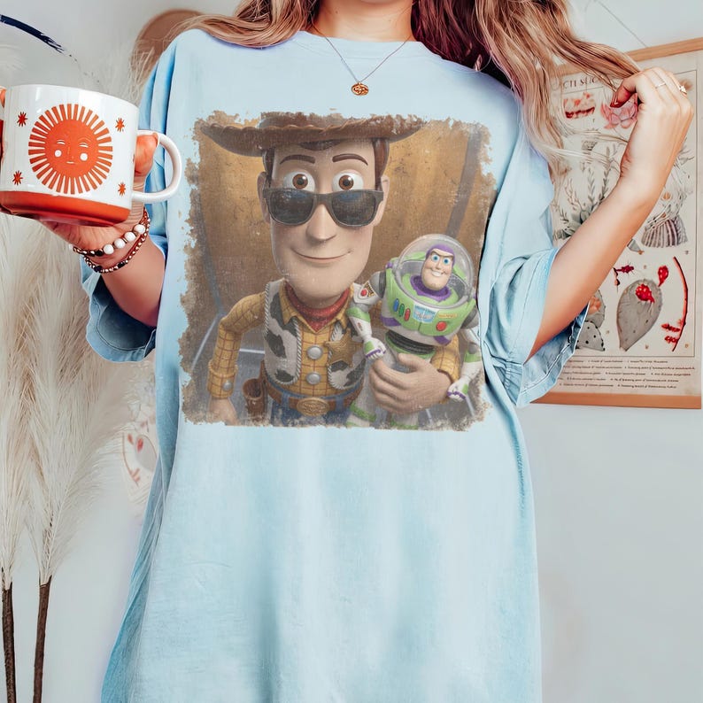 May include: A light blue t-shirt featuring a graphic print of Woody wearing sunglasses and holding Buzz Lightyear. The shirt is paired with a white mug decorated with a large orange sun design and a beaded bracelet on the wrist.
