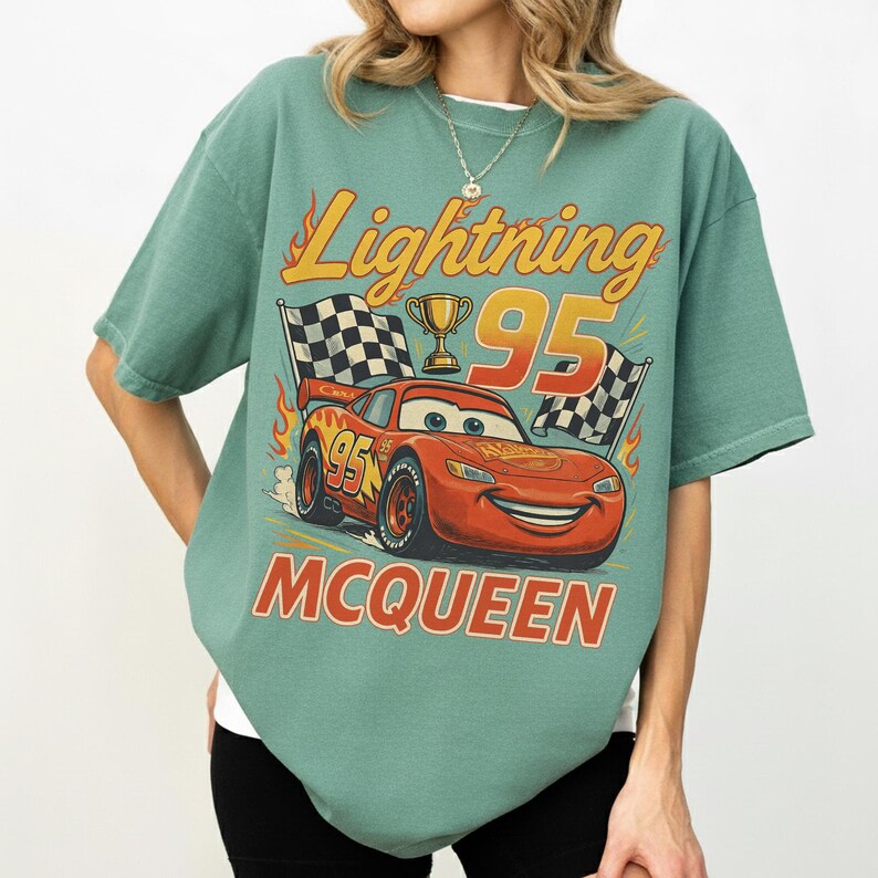 May include: A teal t-shirt with a graphic of Lightning McQueen from the Cars film. The design includes the text "Lightning 95 McQueen", a checkered flag, and a trophy. The shirt has short sleeves and a crew neck.