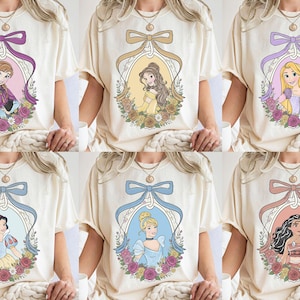 K&ouml;nnte beinhalten: Cremefarbene T-Shirts mit sechs verschiedenen Prinzessinnen-Designs. Jedes Design ist von einem dekorativen Oval mit Schleife und floralen Akzenten umrahmt. Die Prinzessinnen sind Anna, Belle, Rapunzel, Schneewittchen, Aschenputtel und Moana.