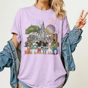 K&ouml;nnte beinhalten: Helllila T-Shirt mit einem farbenfrohen Grafikdesign von Disney-Figuren vor einem Schloss, einem Zug und einer Kugel. Die Grafik zeigt Micky Maus, Minnie Maus, Donald Duck, Daisy Duck, Goofy und Pluto. Feuerwerk im Hintergrund.