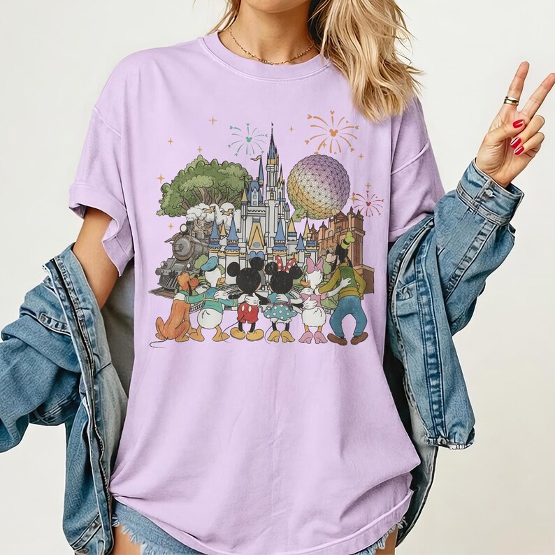 K&ouml;nnte beinhalten: Helllila T-Shirt mit einem farbenfrohen Grafikdesign von Disney-Figuren vor einem Schloss, einem Zug und einer Kugel. Die Grafik zeigt Micky Maus, Minnie Maus, Donald Duck, Daisy Duck, Goofy und Pluto. Feuerwerk im Hintergrund.