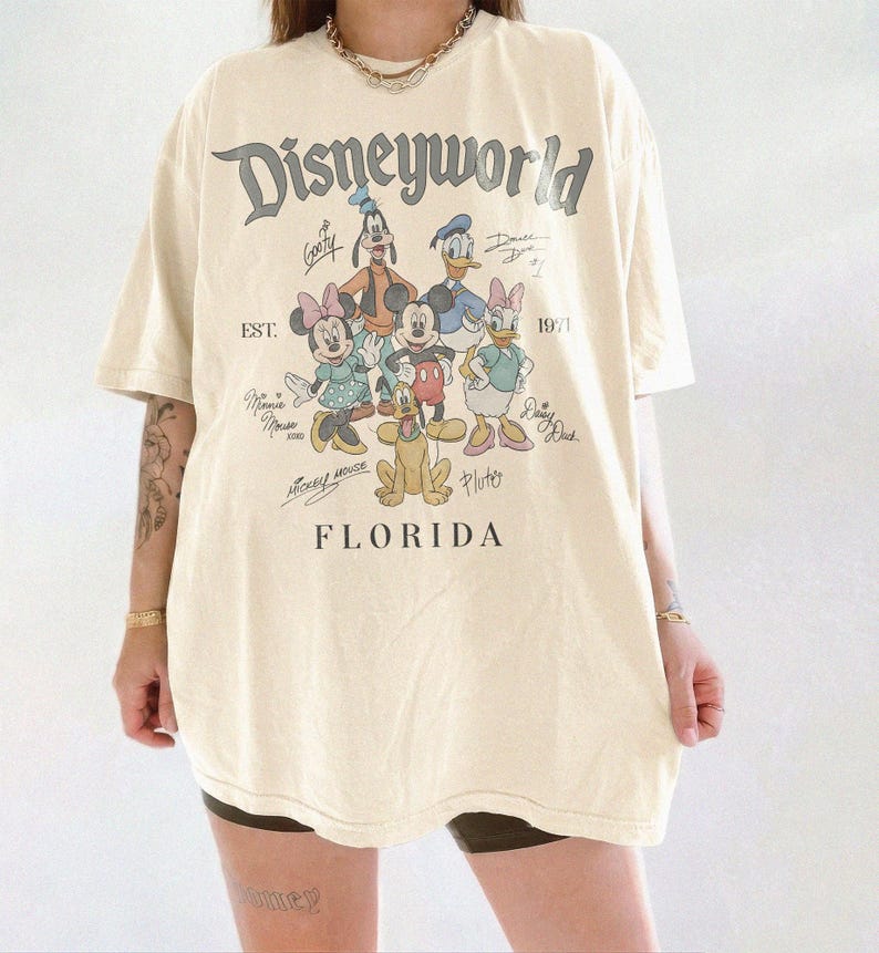 K&ouml;nnte beinhalten: Cremefarbenes &uuml;bergro&szlig;es T-Shirt mit dem Text "Disneyworld" &uuml;ber einer Grafik von Disney-Figuren, darunter Mickey Mouse, Minnie Mouse, Donald Duck und Goofy. Der Text "EST. 1971" und "FLORIDA" sind ebenfalls auf dem Shirt aufgedruckt.