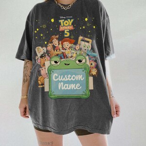 Puede incluir: Camiseta gris oscuro con un gr&aacute;fico colorido de los personajes de Toy Story 5. La camiseta incluye una rana que sostiene un letrero que dice "Custom Name". La camiseta es de corte holgado.