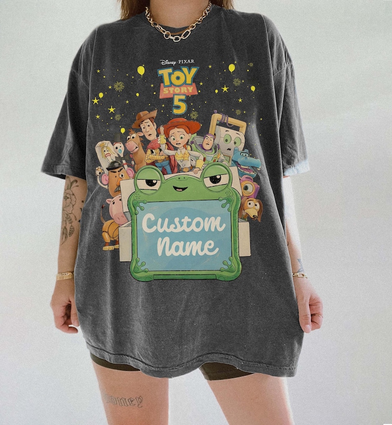 Puede incluir: Camiseta gris oscuro con un gr&aacute;fico colorido de los personajes de Toy Story 5. La camiseta incluye una rana que sostiene un letrero que dice "Custom Name". La camiseta es de corte holgado.