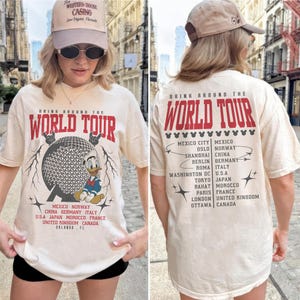 May include: Cream-colored t-shirt with "DRINK AROUND THE WORLD TOUR" in red text. The front features a graphic of a globe and Donald Duck. The back lists cities like Mexico City, Oslo, and Shanghai.