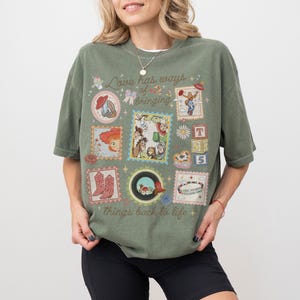 May include: An olive green short-sleeve t-shirt featuring a graphic print with the text "Love has ways of bringing things back to life." The design includes various postage stamp-style frames containing cartoon characters, cowboy boots, and blocks.
