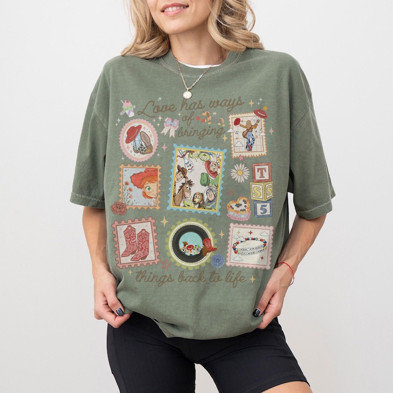 May include: An olive green short-sleeve t-shirt featuring a graphic print with the text "Love has ways of bringing things back to life." The design includes various postage stamp-style frames containing cartoon characters, cowboy boots, and blocks.