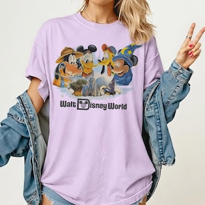 Pu&ograve; includere: T-shirt lilla chiaro con una grafica dei personaggi Disney: Pippo, Paperino, Pluto e Topolino. Il testo "Walt Disney World" &egrave; stampato sotto i personaggi.