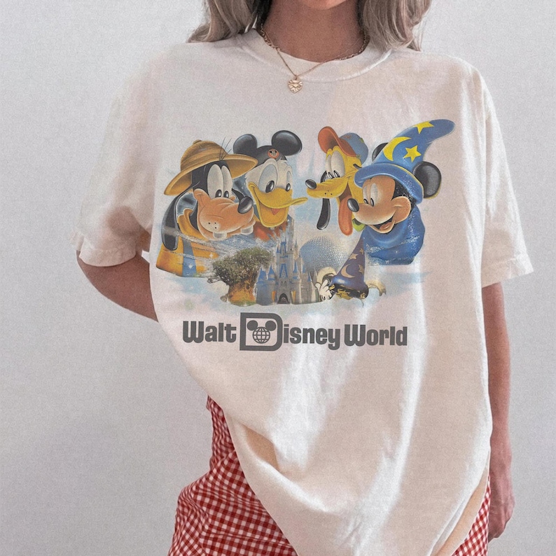 Pu&ograve; includere: T-shirt color crema con una grafica colorata dei personaggi Disney: Pippo, Paperino, Topolino e un castello. Il testo "Walt Disney World" &egrave; stampato sotto in nero.