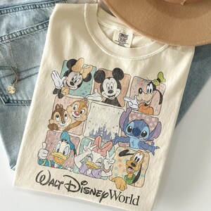 K&ouml;nnte beinhalten: Cremefarbenes T-Shirt mit einem farbenfrohen Aufdruck von Disney-Figuren, darunter Micky Maus, Minnie Maus und Goofy. Auf dem Shirt steht "Walt Disney World". Ein beiger Hut und blaue Jeans sind im Hintergrund.