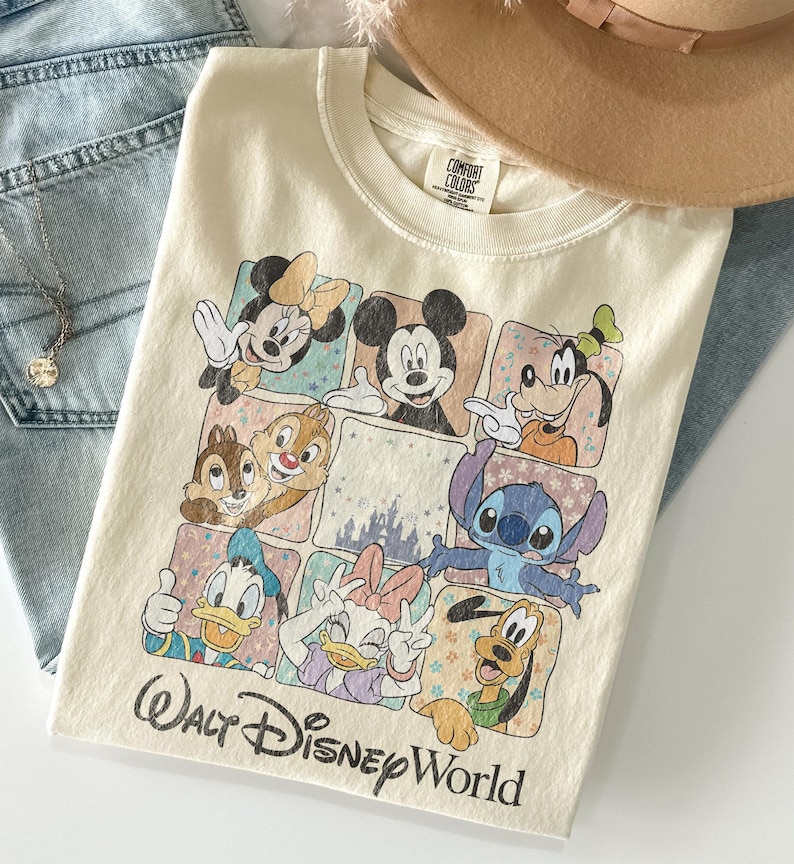 K&ouml;nnte beinhalten: Cremefarbenes T-Shirt mit einem farbenfrohen Aufdruck von Disney-Figuren, darunter Micky Maus, Minnie Maus und Goofy. Auf dem Shirt steht "Walt Disney World". Ein beiger Hut und blaue Jeans sind im Hintergrund.