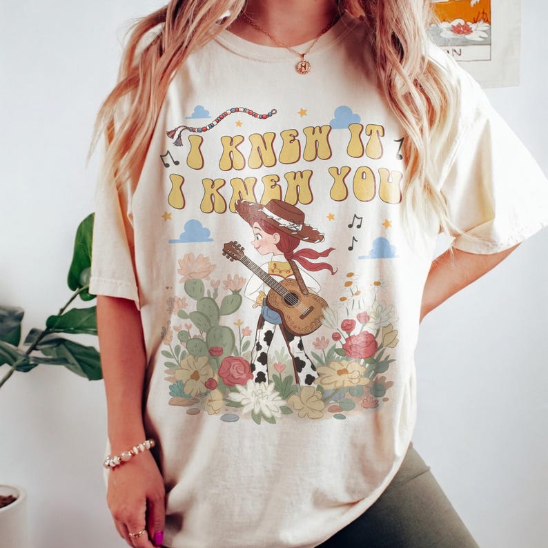 May include: A cream-colored short-sleeve t-shirt featuring a graphic print of a cowgirl playing an acoustic guitar in a desert landscape with cacti and flowers. The text "I KNEW IT I KNEW YOU" is printed in large yellow letters above the character.
