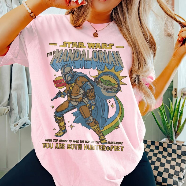 May include: Pink t-shirt featuring a graphic of The Mandalorian and Grogu from Star Wars. The design includes text that reads "The Mandalorian" and "When you choose to walk the way of the Mandalore, you are both hunter & prey."
