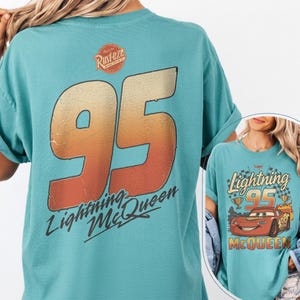 K&ouml;nnte beinhalten: T&uuml;rkisfarbenes T-Shirt mit einer gro&szlig;en 95 in Orange und Gelb, und dem Text "Lightning McQueen". Das Shirt hat auch ein "Rust-eze"-Logo auf dem oberen R&uuml;cken und eine Grafik von Lightning McQueen auf der Vorderseite.