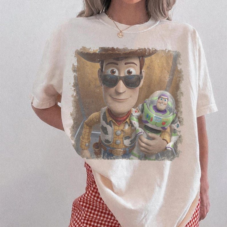 May include: A cream-coloured short-sleeve t-shirt featuring a graphic print of the characters Woody and Buzz Lightyear. Woody is wearing dark sunglasses, and the design has a distressed, textured appearance centred on the front of the shirt.