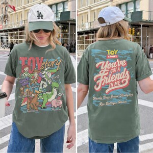 K&ouml;nnte beinhalten: Ein olivgr&uuml;nes T-Shirt mit Toy Story-Grafiken. Die Vorderseite zeigt Jessie, Woody, Buzz Lightyear, Slinky Dog, Bullseye, Rex und Hamm mit dem Text Toy Story Since 1995. Die R&uuml;ckseite zeigt den Text You've Got A Friends In Me.