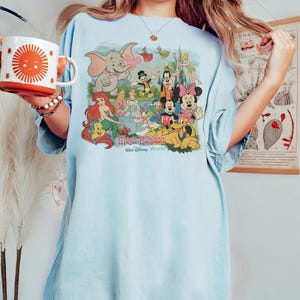 May include: Light blue t-shirt featuring a colorful graphic of Disney characters, including Dumbo, Mickey and Minnie Mouse, and Ariel. The graphic includes the text "Magic Kingdom Walt Disney World". The shirt has short sleeves and a relaxed fit.