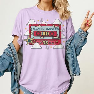 May include: A light purple t-shirt featuring a graphic of a red cassette tape with the text "I KNEW IT, I KNEW YOU" and "TS". The design includes a yellow star with the number "13", white clouds, musical notes, and small yellow stars.