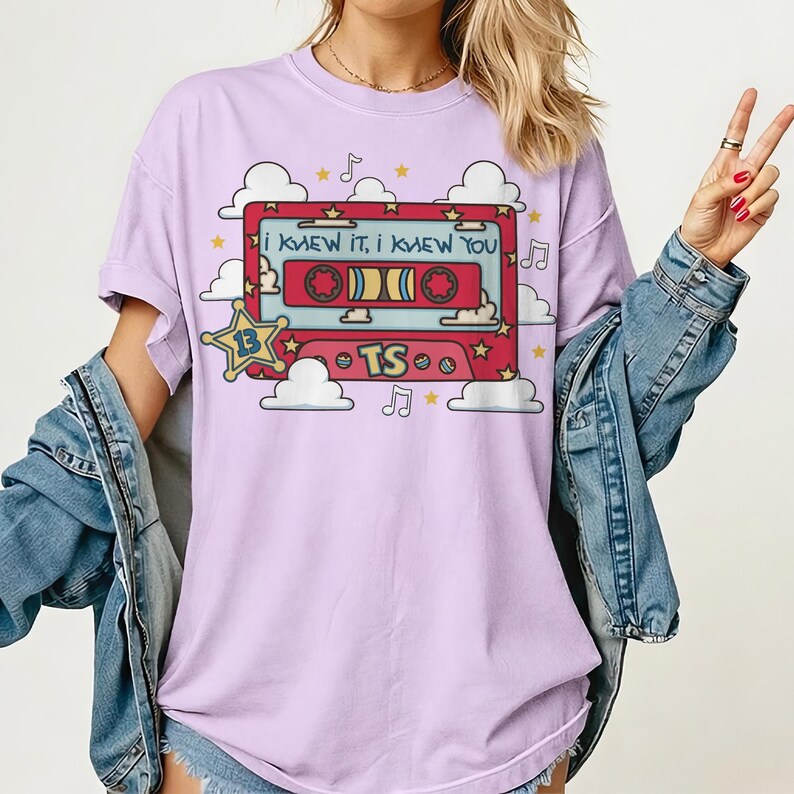 May include: A light purple t-shirt featuring a graphic of a red cassette tape with the text "I KNEW IT, I KNEW YOU" and "TS". The design includes a yellow star with the number "13", white clouds, musical notes, and small yellow stars.