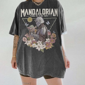 May include: Dark gray t-shirt featuring a graphic of The Mandalorian and Grogu surrounded by flowers. The text "MANDALORIAN - THIS IS THE WAY" is printed above the image in white. The shirt has short sleeves and a relaxed fit.