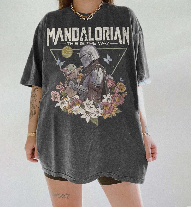May include: Dark gray t-shirt featuring a graphic of The Mandalorian and Grogu surrounded by flowers. The text "MANDALORIAN - THIS IS THE WAY" is printed above the image in white. The shirt has short sleeves and a relaxed fit.