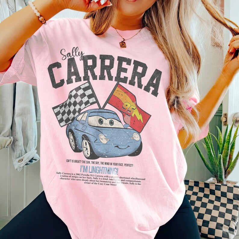May include: Light pink t-shirt featuring a graphic of a blue cartoon car with the text "Sally Carrera" above it. The design includes checkered and red flags, and the text "I'M LINGHTNING!" below the car.