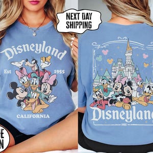 May include: A light blue t-shirt featuring a Disneyland graphic with Mickey Mouse, Minnie Mouse, Donald Duck, and other cartoon characters. The front of the shirt has the Disneyland logo and the text "Est 1955 California". The back of the shirt has a castle graphic.