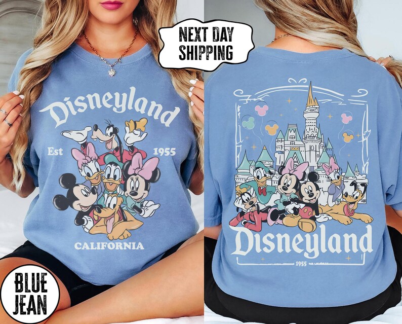 May include: A light blue t-shirt featuring a Disneyland graphic with Mickey Mouse, Minnie Mouse, Donald Duck, and other cartoon characters. The front of the shirt has the Disneyland logo and the text "Est 1955 California". The back of the shirt has a castle graphic.