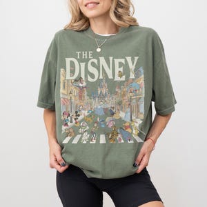 May include: Sage green t-shirt with the text "THE DISNEY" and a colourful illustration of Disney characters crossing a street. The graphic includes Mickey Mouse, Cinderella, and other beloved characters.
