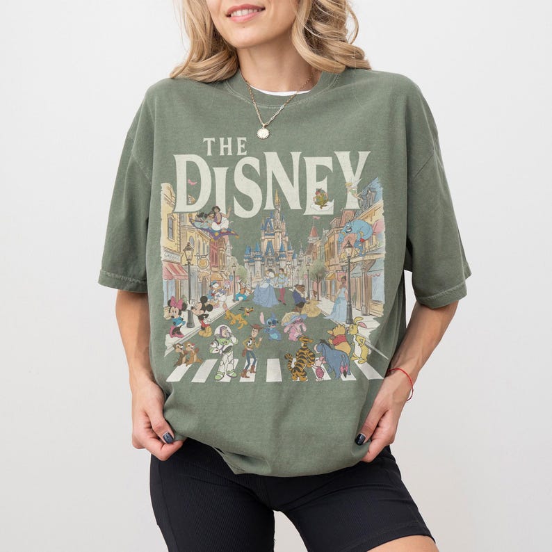 May include: Sage green t-shirt with the text "THE DISNEY" and a colourful illustration of Disney characters crossing a street. The graphic includes Mickey Mouse, Cinderella, and other beloved characters.