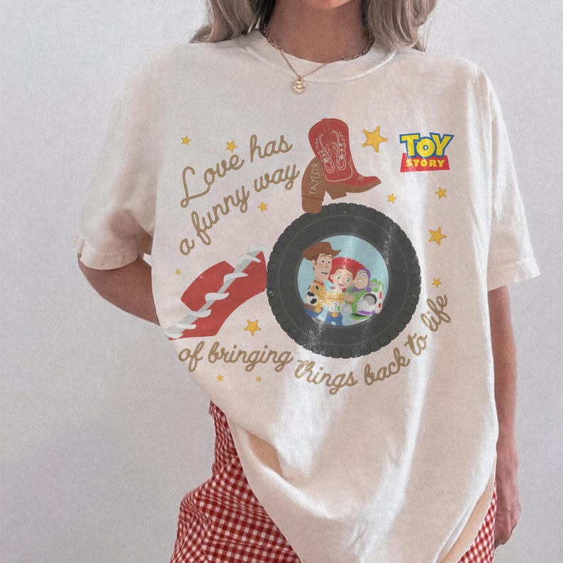 May include: A cream-colored short-sleeve t-shirt featuring a Toy Story graphic. The design includes a red cowboy boot, a red hat, and a black tire framing Woody, Jessie, and Buzz Lightyear. Text reads: Love has a funny way of bringing things back to life.