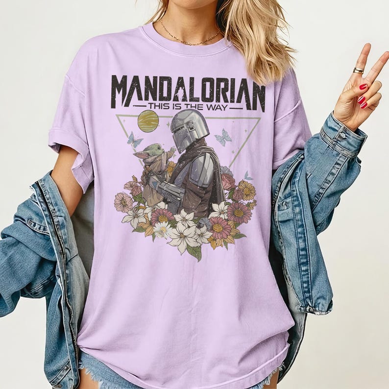 May include: A light purple t-shirt featuring a graphic of The Mandalorian and Grogu surrounded by flowers and butterflies. The text "MANDALORIAN - THIS IS THE WAY" is printed across the top of the design.