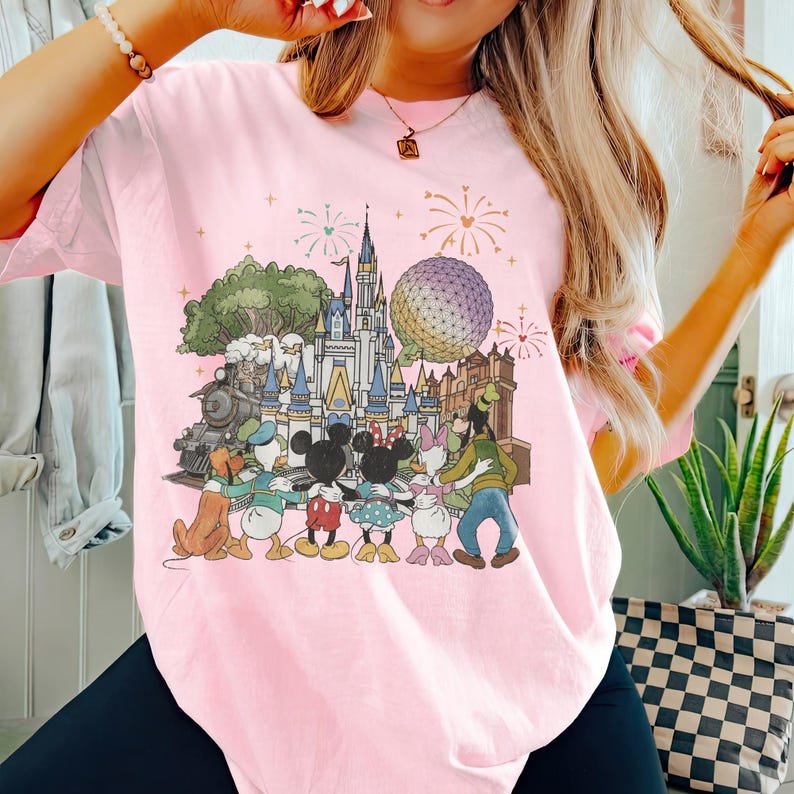 K&ouml;nnte beinhalten: Hellrosa T-Shirt mit einem farbenfrohen Aufdruck von Disney-Figuren und Wahrzeichen, darunter ein Schloss, Spaceship Earth und ein Zug. Das Design umfasst Micky und Minnie Maus, Donald und Daisy Duck und Goofy. Feuerwerk ist ebenfalls abgebildet.
