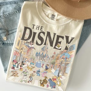 May include: Cream-coloured t-shirt with the text "THE DISNEY" and a colourful illustration of Disney characters and a street scene. The shirt is folded on top of blue jeans and a tan hat.