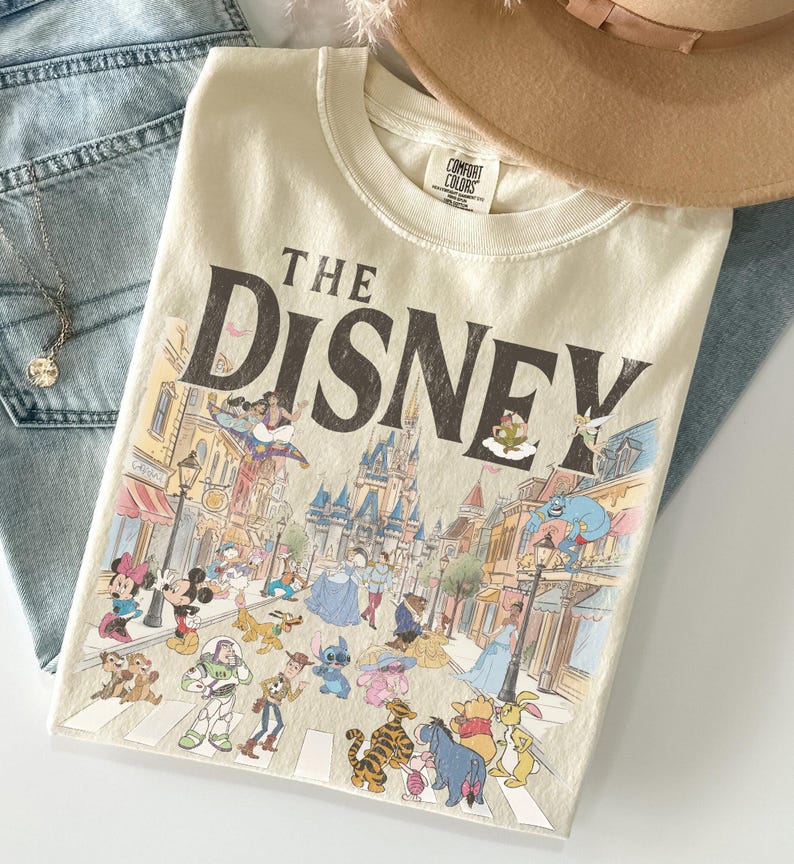 May include: Cream-coloured t-shirt with the text "THE DISNEY" and a colourful illustration of Disney characters and a street scene. The shirt is folded on top of blue jeans and a tan hat.