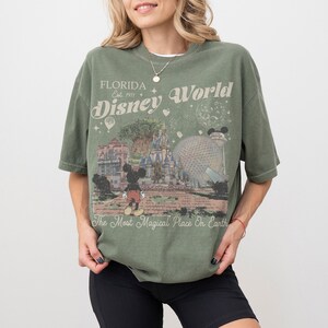 Puede incluir: Camiseta verde oliva con un dise&ntilde;o de Disney World en Florida. El dise&ntilde;o incluye a Mickey Mouse, el castillo de Cenicienta y el texto "Florida Est. 1971 Disney World" y "The Most Magical Place On Earth."