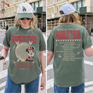 May include: Olive green t-shirt with a graphic featuring Minnie Mouse, a globe, and the text "DRINK AROUND THE WORLD TOUR." The back of the shirt lists various cities. The shirt is paired with blue jeans and a white baseball cap.