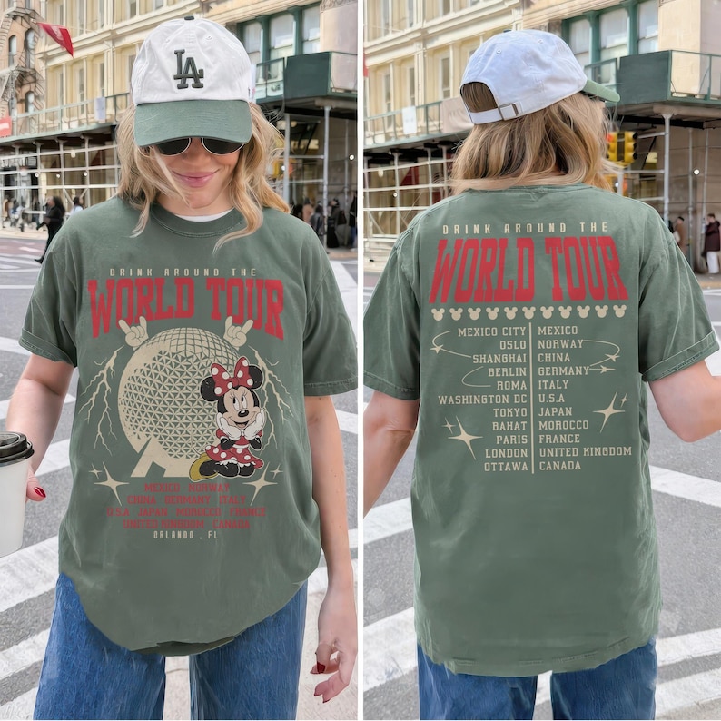 May include: Olive green t-shirt with a graphic featuring Minnie Mouse, a globe, and the text "DRINK AROUND THE WORLD TOUR." The back of the shirt lists various cities. The shirt is paired with blue jeans and a white baseball cap.