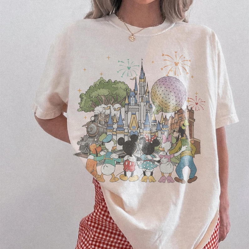 K&ouml;nnte beinhalten: Cremefarbenes T-Shirt mit einer farbenfrohen Illustration eines Themenparks mit Schloss, Zug und Figuren wie Mickey und Minnie Mouse. Feuerwerk und eine gro&szlig;e Kugel sind ebenfalls im Design dargestellt.
