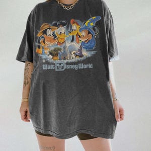 Pu&ograve; includere: T-shirt grigio scuro con una grafica dei personaggi Disney: Pippo, Paperino, Pluto e Topolino con un cappello da mago. Il testo "Walt Disney World" &egrave; stampato sotto. La maglietta ha maniche corte e una vestibilit&agrave; ampia.