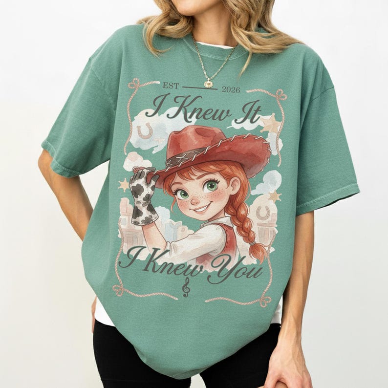 May include: A sage green t-shirt featuring a graphic of a cartoon character wearing a brown cowboy hat and a cow-print glove. The text reads EST 2026, I Knew It, and I Knew You, surrounded by illustrations of stars, clouds, and horseshoes.