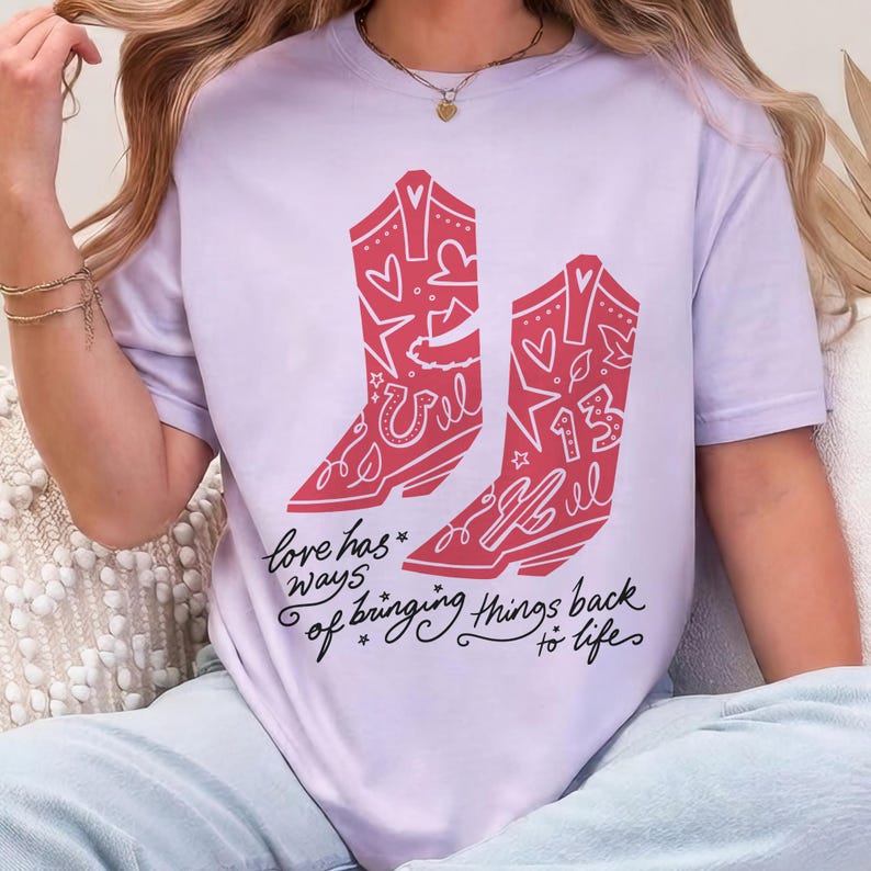 K&ouml;nnte beinhalten: Ein hellviolettes T-Shirt mit einer Grafik von zwei roten Cowboystiefeln, die mit Herzen, Sternen und der Zahl 13 verziert sind. Unter den Stiefeln steht in schwarzer Schreibschrift: love has ways of bringing things back to life, mit kleinen Sternen.