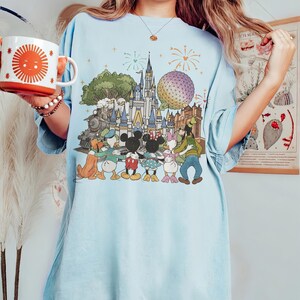 K&ouml;nnte beinhalten: Hellblaues T-Shirt mit einem farbenfrohen Grafikdesign eines Vergn&uuml;gungsparks mit Schloss, Zug und Zeichentrickfiguren wie Mickey und Minnie Mouse. Das Design zeigt auch Feuerwerk und eine runde Struktur. Eine wei&szlig;e Tasse mit einem orangefarbenen Sonnendesign wird gehalten.