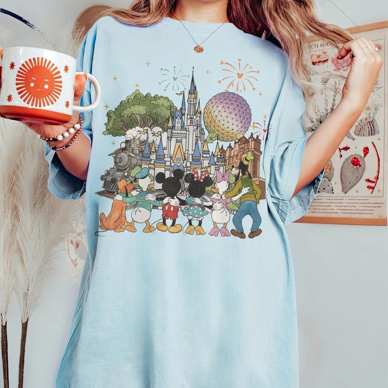 K&ouml;nnte beinhalten: Hellblaues T-Shirt mit einem farbenfrohen Grafikdesign eines Vergn&uuml;gungsparks mit Schloss, Zug und Zeichentrickfiguren wie Mickey und Minnie Mouse. Das Design zeigt auch Feuerwerk und eine runde Struktur. Eine wei&szlig;e Tasse mit einem orangefarbenen Sonnendesign wird gehalten.